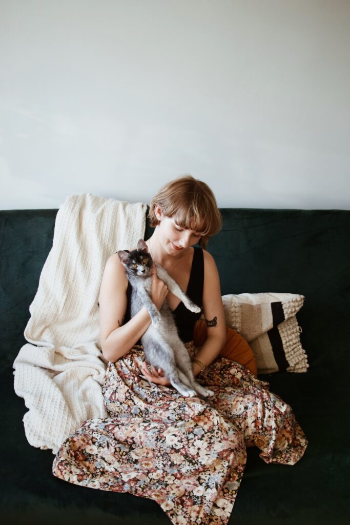 Izabela siedzi na zielonej kanapie i trzyma kotkę Kasjopeję. W tle poduszki i ozdobny koc.