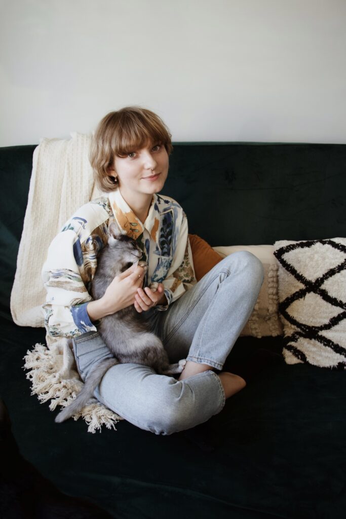 Izabela Stańczyk siedzi na kanapie i trzyma kotkę Kasjopeję na rękach. Kotka dostaje przysmak z tubki.