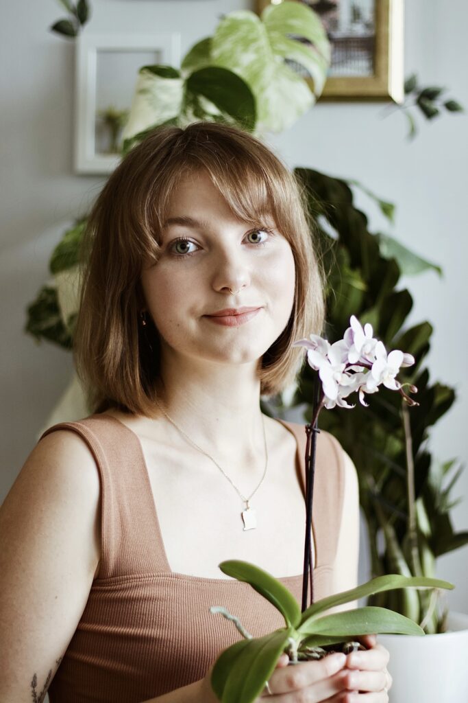 Izabela Stańczyk pozuje do zdjęcia trzymając w dłoniach storczyka. W le monstera variegata i zamioculcas supernova.